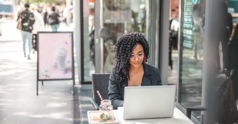 High angle of pensive African American female freelancer in glasses and casual clothes focusing on screen and interacting with netbook while sitting at table with glass of yummy drink on cafe terrace in sunny day